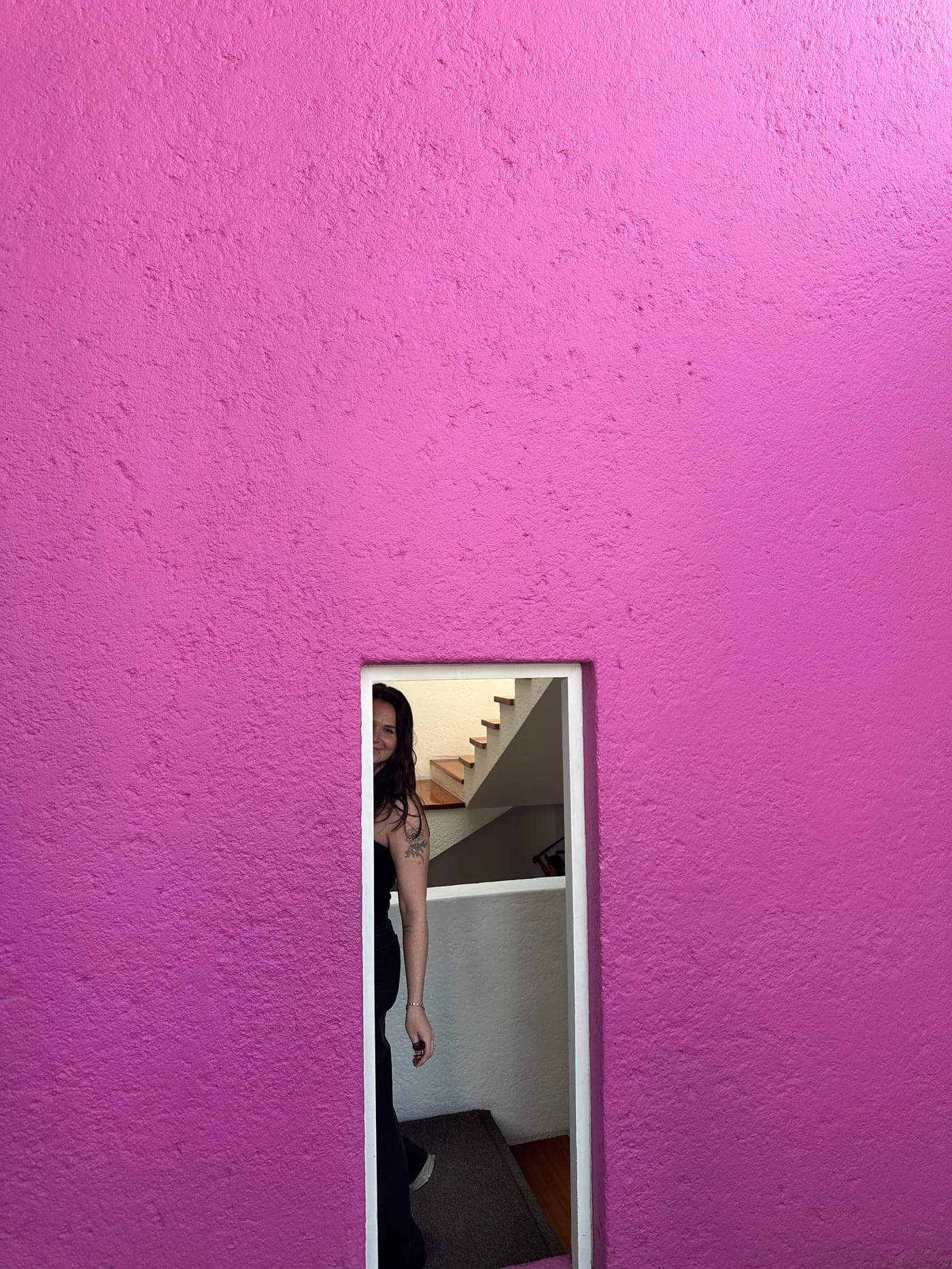 Liz Fryett photographed through a narrow window cutout in a pink stairwell wall.